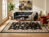 Load image into Gallery viewer, A large traditional-style area rug with a black field and ornate beige floral border, styled in a bright living room with a dark gray sofa, wooden coffee table, red armchair, and potted plant.