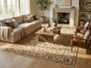 Load image into Gallery viewer, A large traditional-style area rug with an intricate floral pattern in beige, brown, and muted blue tones, styled in a bright, modern living room with a beige sofa, wooden coffee table, and fireplace.