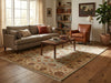 Load image into Gallery viewer, A large traditional-style area rug with a beige, brown, and muted red floral medallion pattern, shown in a styled living room with a gray sofa, brown leather armchair, wooden coffee table, and hardwood floors.