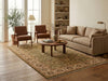 Load image into Gallery viewer, A large traditional-style area rug in beige, green, and brown tones is styled in a warm, modern living room with a beige sofa, two brown armchairs, a round wooden coffee table, and a floor lamp.
