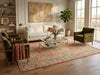 Load image into Gallery viewer, A styled living room featuring a large rectangular area rug with a distressed floral pattern in muted pink, beige, and green tones. The rug is placed on a light wood floor in front of a cream sofa, flanked by two olive green armchairs with a glass coffee table in the center.