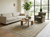 Load image into Gallery viewer, A styled living room scene featuring a light beige three-seater sofa, a matching armchair, a round wooden coffee table, and a large patterned area rug on a light wood floor. A tall potted fiddle-leaf fig plant stands near a large black-framed window.