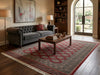 Load image into Gallery viewer, A large traditional area rug with a red and gray geometric pattern and white fringe, styled in a warm, classic living room setting with a gray tufted sofa and a wooden coffee table.