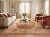 Load image into Gallery viewer, A styled living room featuring a large traditional area rug with a central medallion pattern in beige, rust, and pink. The room includes a cream sofa with accent pillows, two rust-colored armchairs, a round wooden coffee table with a floral arrangement, and a white floor lamp.