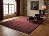 Load image into Gallery viewer, A large traditional area rug with a deep red and brown pattern, styled in a warm, classic living room with a dark leather armchair, matching ottoman, wooden floor, and built-in shelves.