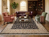 Load image into Gallery viewer, A styled living room featuring a large traditional oriental area rug with a black, beige, and pink floral pattern. The rug is styled in a warm, classic room with a dark brown leather Chesterfield sofa, a dusty pink armchair, a sage green armchair, and a glass coffee table.