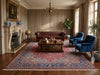 Load image into Gallery viewer, A large traditional oriental area rug with a red, blue, and beige pattern is styled in a classic, elegant living room with a brown leather Chesterfield sofa, blue velvet armchairs, a wooden coffee table, and a fireplace.