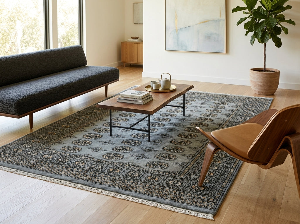 A large traditional-style area rug in muted blue-gray and beige tones, styled in a bright modern living room with a dark gray sofa, wooden coffee table, and a brown wooden lounge chair.