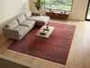 Load image into Gallery viewer, A large red Persian-style area rug with intricate circular patterns and cream fringe, styled in a modern living room with a beige sectional sofa and a wooden coffee table.